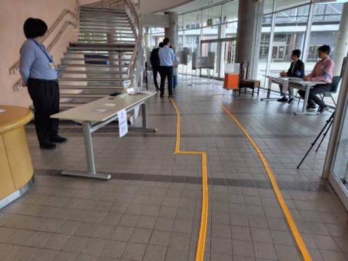 大学講堂のロビーを使用した障がい者対応の模擬投票所の様子。 ココテープ(視覚障害者誘導用ブロック)で入口から投票台、投票箱へのガイドが引いてあります。
