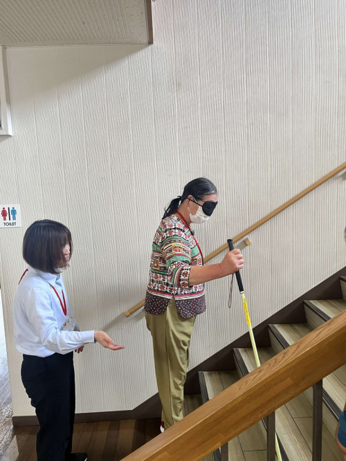 アイマスクをして白杖を持ち階段を上る体験をしている参加者と、上り方の説明をしている職員