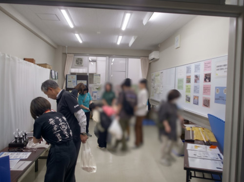 会場となった部屋の全体の様子。 職員が来場者に機器の説明を行っている様子。